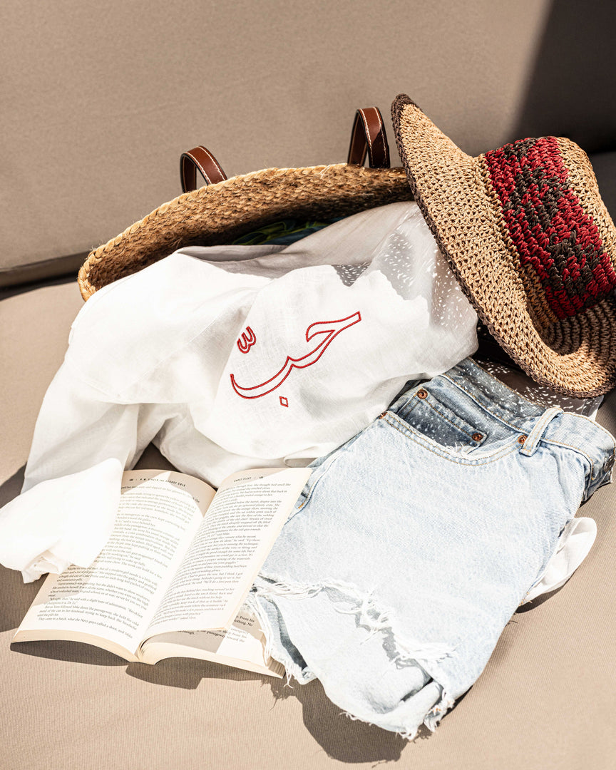 Clothing items including a white shirt with Arabic text, denim shorts, and a straw hat on a beige surface.
