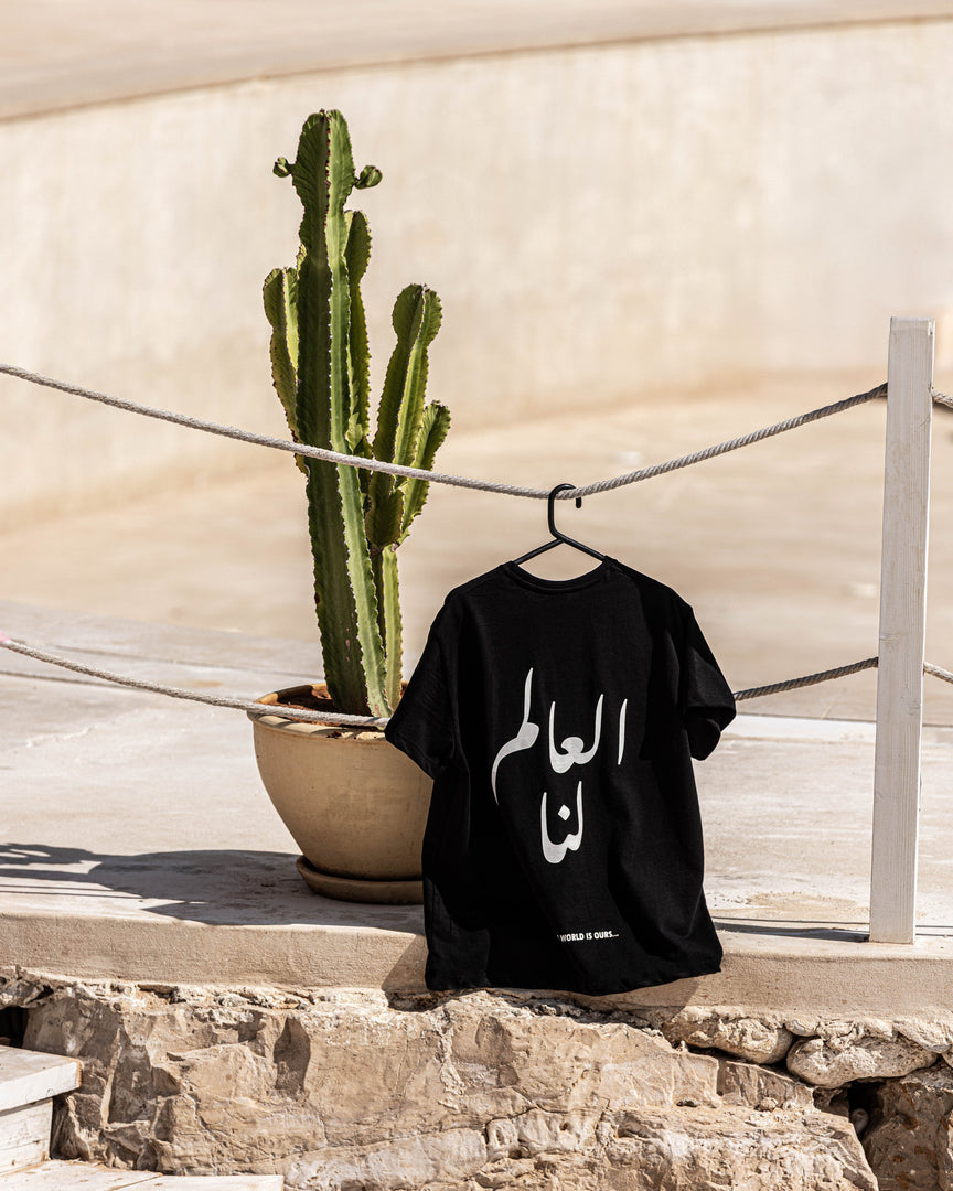 Black t-shirt with Arabic text on a hanger next to a cactus plant outdoors.