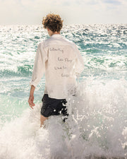Person standing in ocean waves wearing a white shirt with text.